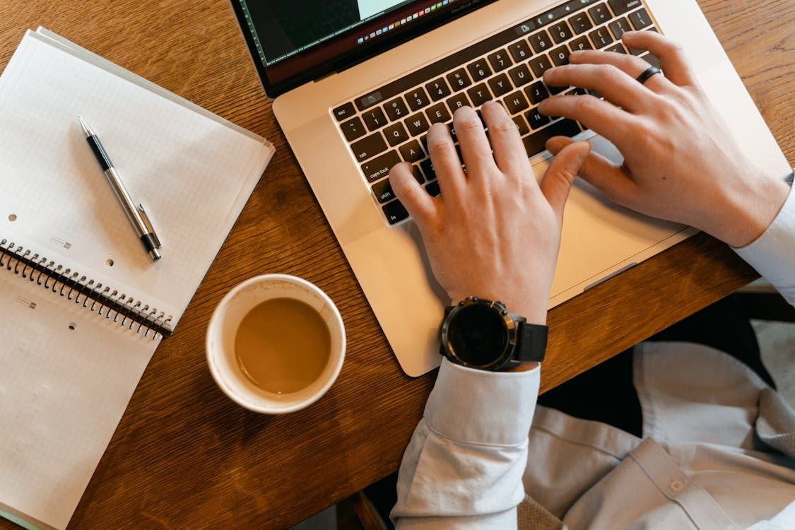 student using a laptop to email a professor at a desk with notebook and coffee
